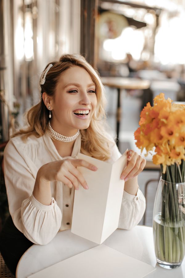 joyful-curly-blonde-lady-stylish-white-blouse-pearl-necklace-smiles-sincerely-sits-street-cafe-by-little-table-with-yellow-bouquet-holds-menu_result